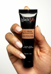 Black Up CC-Creme Multi-Action in einer schwarzen Tube mit mattem Finish und einem beigen Etikett; wird von einer Hand mit sanftem, neutralem Nagellack gehalten.