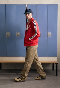 Red zip-up jacket with black and white stripes, beige pants, and yellow shoes with black stripes. Locker background with blue doors.