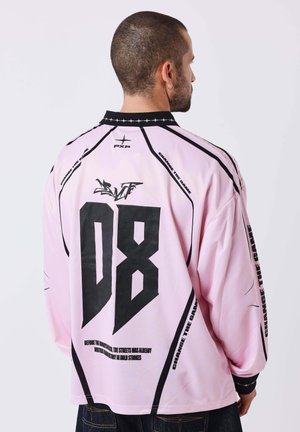 Mann mit kurzen Haaren, der ein pinkfarbenes Langarmshirt mit schwarzem "D8"- und "Change the Game"-Schriftzug auf dem Rücken vor schlichtem Hintergrund trägt.