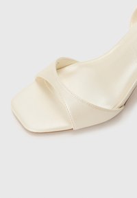 Beige satinsandal med hæl og firkantet tå, med krydsspenner og en glat overflade. Enkel design uten synlig maskinvare.