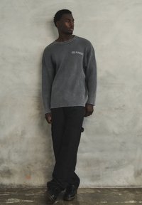 Maglione grigio a coste con scollatura ampia, abbinato a pantaloni cargo neri e scarpe nere. Ricamo distintivo sulla parte frontale del maglione.