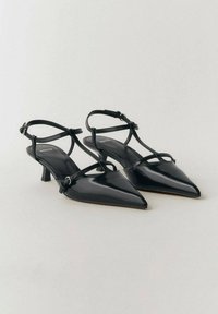Czarne lakierowane szpilki slingback z wąskimi paskami na kostkę, szpiczastymi noskami i małym, blokowym obcasem. Charakteryzują się minimalistycznym stylem.