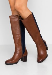 Botas altas hasta la rodilla de cuero marrón y ante azul marino con tacón grueso, que cuentan con una cremallera lateral y suela texturizada.