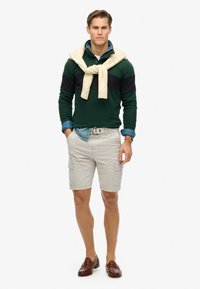 Grøn og sort langærmet polo, blå denimskjorte under, cremefarvet sweater bundet om halsen, beige cargo shorts, brune loafers.