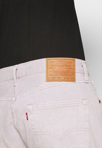 Vaqueros Levi's de color rosa claro combinados con una camiseta negra, mostrando el bolsillo trasero y la etiqueta de cuero color marrón con detalles de talla.