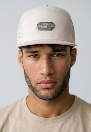 Young man with a short beard wearing a light beige cap with "BLACKSKIES PRO" logo and matching beige shirt, facing forward with neutral expression.