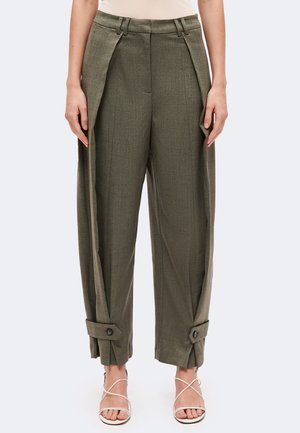 Un mannequin porte un pantalon olive à jambes larges, avec des plis sur le devant et des revers boutonnés aux chevilles, associé à des sandales blanches à brides.