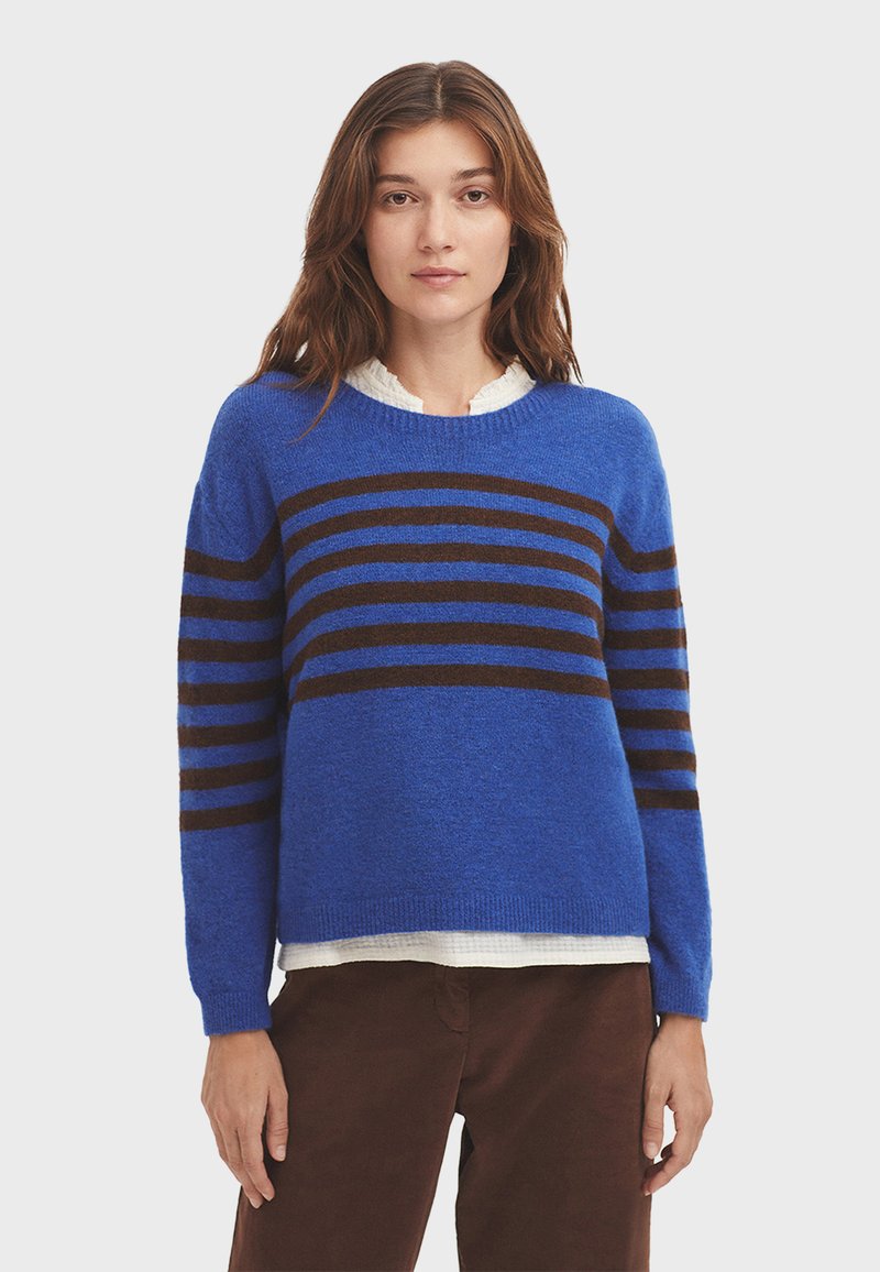 Blaues Sweatshirt aus weichmaterial mit braunen horizontalen Streifen. Hat einen Rundhalsausschnitt und lange Ärmel. Über einem weißen Oberteil getragen.