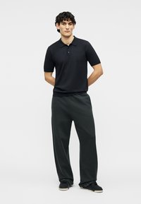 Νεαρός άνδρας που στέκεται με τα χέρια πίσω από την πλάτη, φορώντας μαύρο πόλο με κοντά μανίκια, σκουρόχρωμο φαρδύ παντελόνι και μαύρα αθλητικά παπούτσια, σε λευκό φόντο.