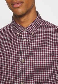 Homme portant une chemise à carreaux rouge, blanche et bleue avec un col boutonné, visible du cou à la poitrine.