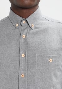 Grijze button-up shirt van gestructureerde stof, met een puntige kraag, zes knopen en een enkele borstzak. Lichtbruine knopen bieden contrast.