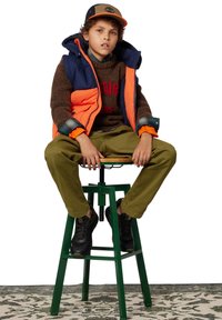 Gilet imbottito blu navy e arancione, maglione in maglia marrone con testo rosso, chinos verdi, sneaker nere e un cappellino marrone/nero con logo. Seduto su uno sgabello verde.
