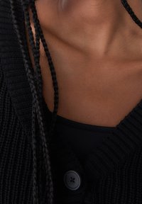 Černý žebrovaný cardigan s hlubokým V-výstřihem, který má velké zapínání na knoflík. Textura je pletená a pod ním je vidět černý top.