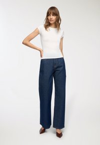 Biała dopasowana koszulka z krótkim rękawem w zestawieniu z wysokimi, szerokimi niebieskimi dżinsami. Gładki materiał, jasnoniebieskie przeszycia i spiczaste brązowe buty.