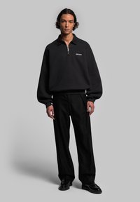 Sweatshirt noir zippé avec col, présentant une coupe décontractée et des poignets côtelés. Associe à un pantalon noir ample et des chaussures noires brillantes.