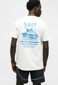 Άνδρας που φοράει λευκό T-shirt με μπλε γραφικό κτήριο και κείμενο «OBEY House of Subversion» που απαριθμεί μεγάλες πόλεις του κόσμου στην πλάτη.