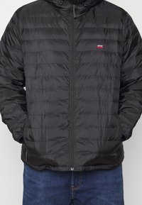 Svart quiltad Levi's-jacka med dragkedja framtill, händerna i fickorna, bärs över blå jeans, visas från nacke till höfter.