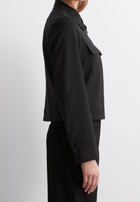 Zwarte cropped jas met een kraag, borstzak, lange mouwen en manchetten met knopen. Gemaakt van een glad materiaal met een matte afwerking.