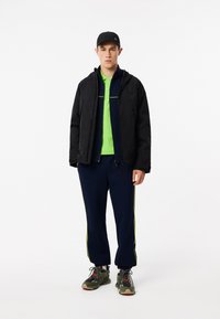 Giacca nera, polo verde, pantaloni blu navy con strisce neon, sneakers grigie. Design casual e sportivo, con texture e materiali vari.