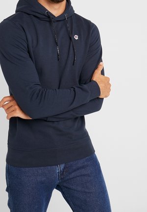 Hombre con los brazos cruzados, vistiendo una sudadera con capucha azul marino con cordones y un pequeño logo en el pecho, combinado con vaqueros azul oscuro, sobre un fondo gris claro.