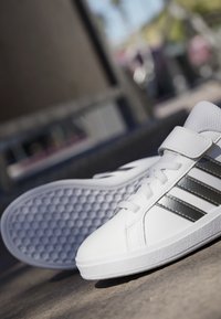 Bijele atletske cipele s glatkom kožnom gornjom stranom, tri metalno crne pruge, klasičnim vezicama i teksturiranom gumiranom potplatom.