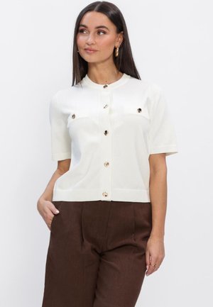 Vrouw met lang donker haar, een wit kort mouwtjes gebreid vest met knopen en bruine hoge taille broek, met haar hand in haar zak, kijkt naar de zijkant.