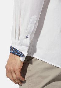 Weißes Langarmhemd mit einem Hahnentritt-Muster an den Ärmeln in Blau, Braun und Weiß. Besitzt einen Knopf und einen schlichten Kragen.