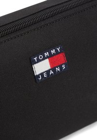 Μαύρο πορτοφόλι από ύφασμα με ραμμένη ετικέτα "TOMMY JEANS" σε ναυτικό μπλε με κόκκινες και λευκές λεπτομέρειες, που παρουσιάζει υφή στην επιφάνεια και λεπτομέρεια με φερμουάρ.
