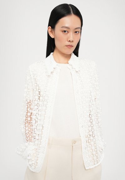 White lace jacket with a floral pattern, featuring a collared neckline and button closure. Worn over a light-colored top and beige trousers.