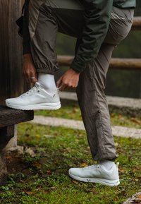 Zapatillas deportivas blancas con una textura suave y un logo, combinadas con pantalones grises ajustados y calcetines blancos. Césped y un banco de madera al fondo.
