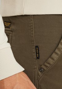 Olive green pants with a textured finish, featuring a visible button and a black label with yellow text, paired with a light beige ribbed cuff.