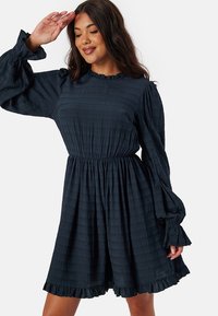 Robe texturée bleu marine avec de longues manches bouffantes et un ourlet froncé. Présente une taille ajustée et un col rond.