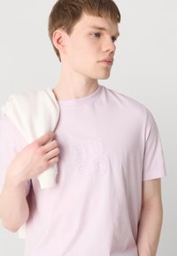 Lyslilla bomulds t-shirt med broderet "HUGO" logo. Model, der holder en hvid sweatshirt over den ene skulder. Korte ærmer, rund halsudskæring.