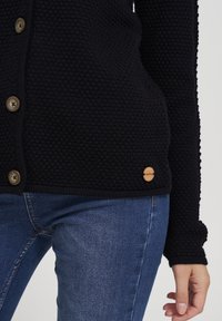 Námořnický pletený cardigan s texturovaným vzorem, vybavený hnědými knoflíky a dřevěným akcentem s logem. Spárováno s modrými džínami.