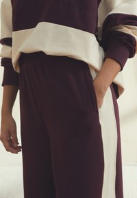 Bordo trenirka z zelnim trakom. Mehka, teksturirana tkanina z udobnim krojem. Vsebuje rebrasti pas in stranske žepe.