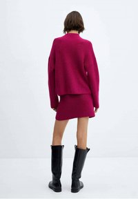 Fuksijaste pletene pulover in ujemajoča krila z ohlapnim krojem, teksturirana tkanina, v kombinaciji z visokimi črnimi škornji. Zadnji pogled proti enotni ozadju.