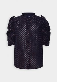 Blouse bleu marine boutonnée avec des pois métalliques rose doré, manches trois-quarts bouffantes et petit col montant, posée à plat.