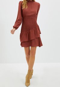 Rostrote Kleid mit langen Ärmeln, gerafftem Taillenbund und geschichteten Rüschen. Glatter Stoff mit hohem Kragen, kombiniert mit beigen Knöchelstiefeln.