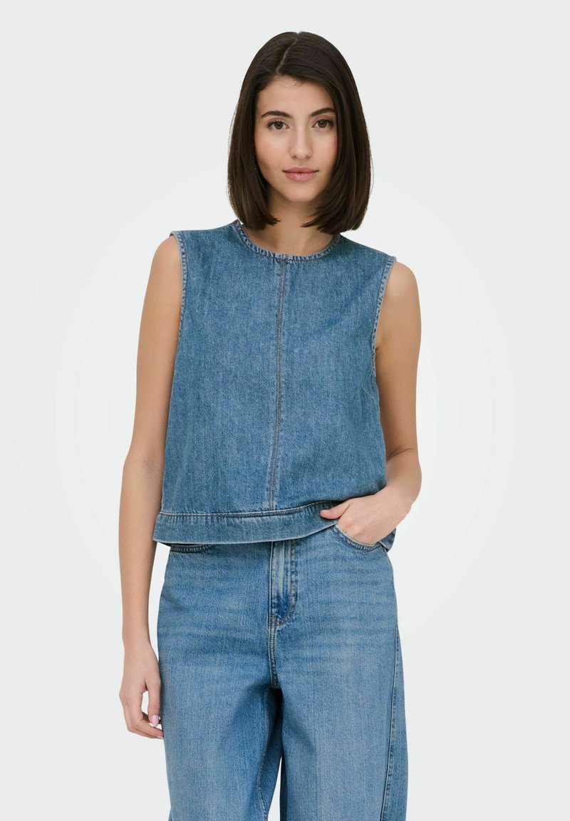 Femme aux cheveux foncés mi-longs, portant un top en jean sans manches et un jean taille haute, main dans la poche, fond neutre.