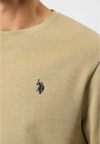 Beige katoenen sweatshirt met een ronde hals. Voorzien van een klein marineblauw geborduurd logo op de borst, zichtbaar textuur in de stof.
