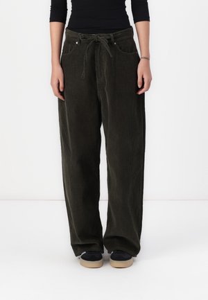 Personne portant un pantalon large en velours côtelé vert foncé avec une ceinture nouée à la taille et des chaussures noires à semelles beiges, se tenant devant un fond blanc.