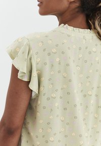 Helles grünes Oberteil mit kurzen Volantsärmeln, strukturiertem Stoff und floralem Muster in Gelb- und Lilatönen. Dezenter Rüschensaum am Ausschnitt.