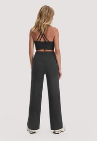 Frau von hinten, die einen schwarzen Sport-BH mit Riemchen, hochtaillierte, weit geschnittene schwarze Hosen und weiße Turnschuhe vor einem einfarbigen Hintergrund trägt.