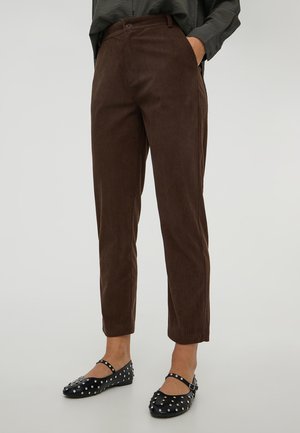 Persona che indossa pantaloni in velluto a coste marroni e scarpe Mary Jane nere con borchie, in piedi con le mani in tasca su uno sfondo semplice.