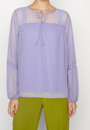 Femme portant un chemisier transparent lavande à manches longues, avec une texture à pois et un lien au niveau du col, associé à un pantalon vert olive.
