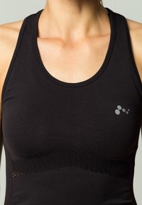 Camiseta sin mangas deportiva negra con escote redondo, tira de malla texturizada en la cintura y un sutil detalle del logo gris en el lado izquierdo.