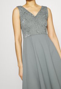 Harmaa mekko, jossa on pitsinen yläosa kukkakuviolla, V-pääntie ja liehuva helma, tehty kevyestä kankaasta.