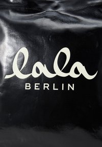 Crna sjajna torba glatke teksture s bijelim, kurzivnim logotipom "lala" iznad riječi "BERLIN" u podebljanom, sans-serif fontu.