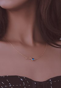 Guld kædelningshalskæde med et kursiv "kærlighed" design og en hjerteformet blå ædelsten akcent. Enkel, elegant og delikat håndværk.