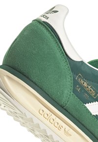 Zapatilla de ante verde y textil con acentos blancos, que presenta una suela texturizada y un logo metálico dorado. Diseño clásico con un toque retro.